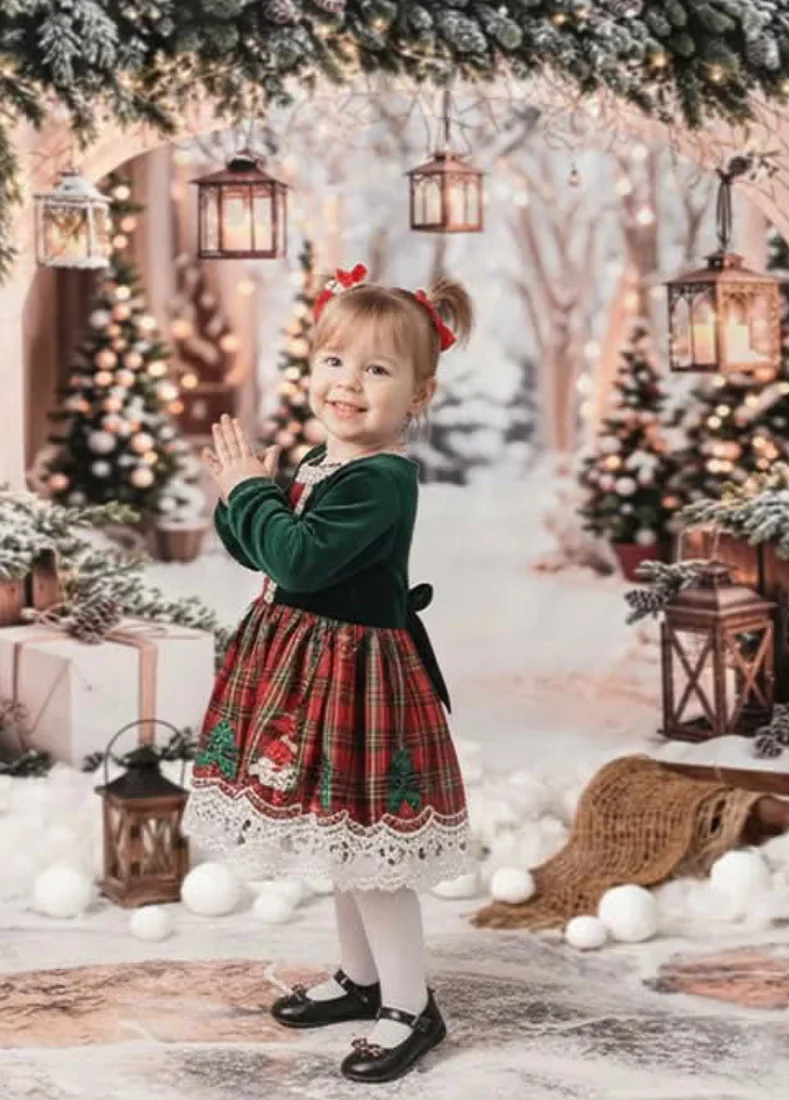 A little girl in a green velvet jacket and tartan dress clapping in a snowy Christmas set
