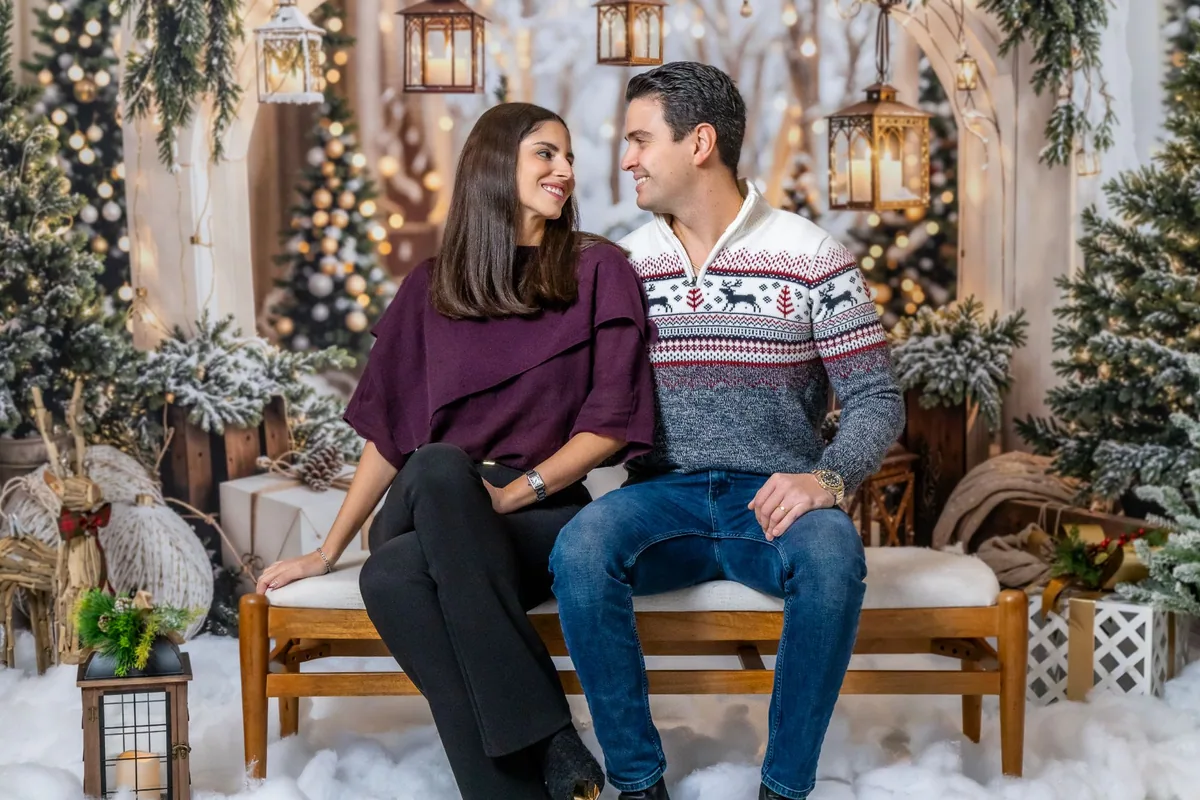 Couple in a burgundy top and a reindeer sweater looking at each other on a bench in the snowy Christmas set